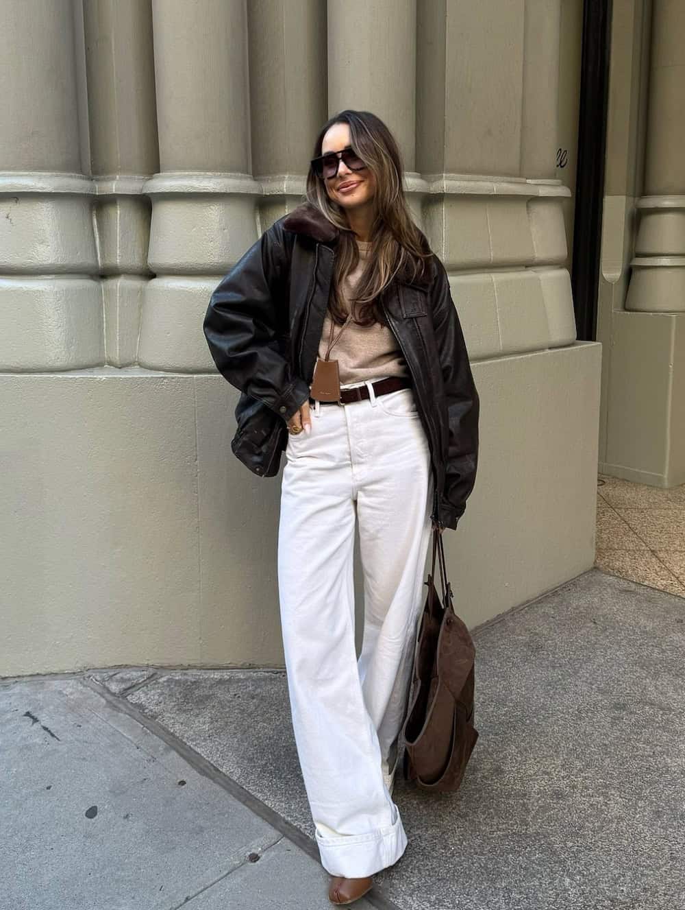 Woman wearing white wide leg jeans with a tan sweater and a black jacket.